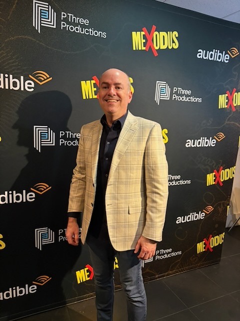 Brian Guy smiling on the Mexodus red carpet in front of a step-and-repeat with Mexodus, Audible, and P Three Productions logos