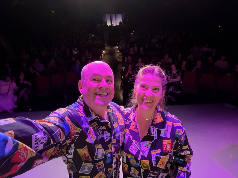 Brian Guy and co-host Josi Twigt on stage in matching pajamas printed with theatre program covers, taking a selfie with the audience at Tacoma Little Theatre behind them