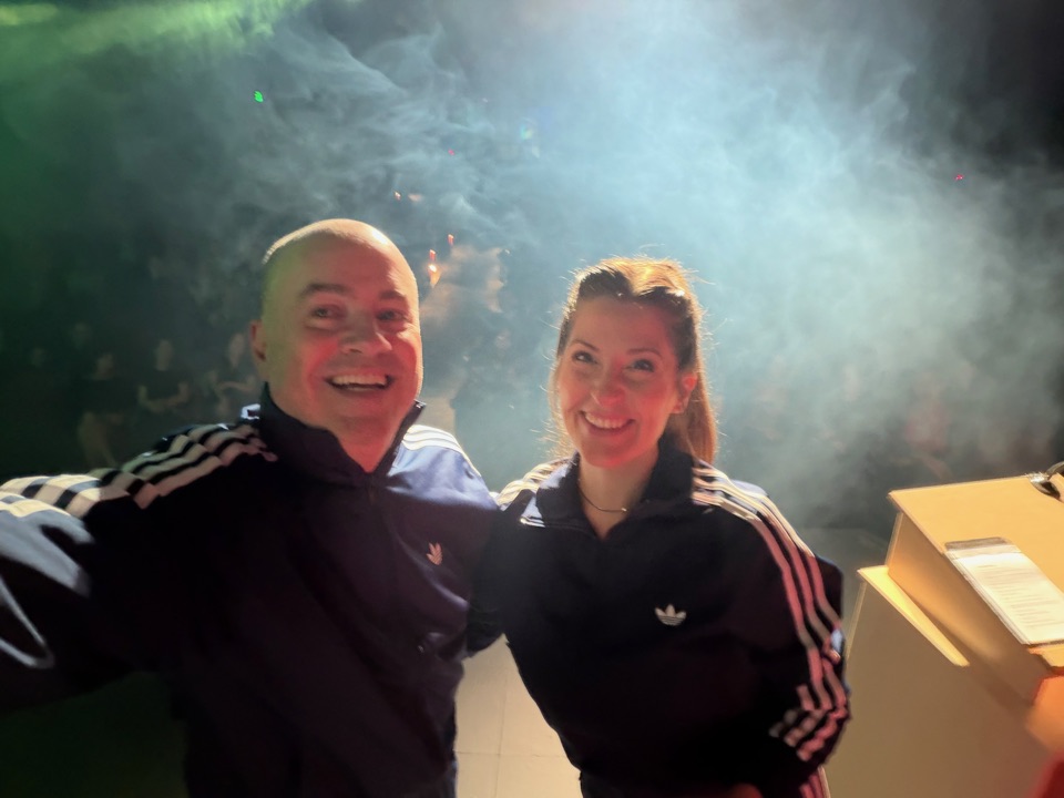 Brian Guy and co-host Josi Twigt on stage in matching navy tracksuits, smiling for a selfie with stage haze behind them